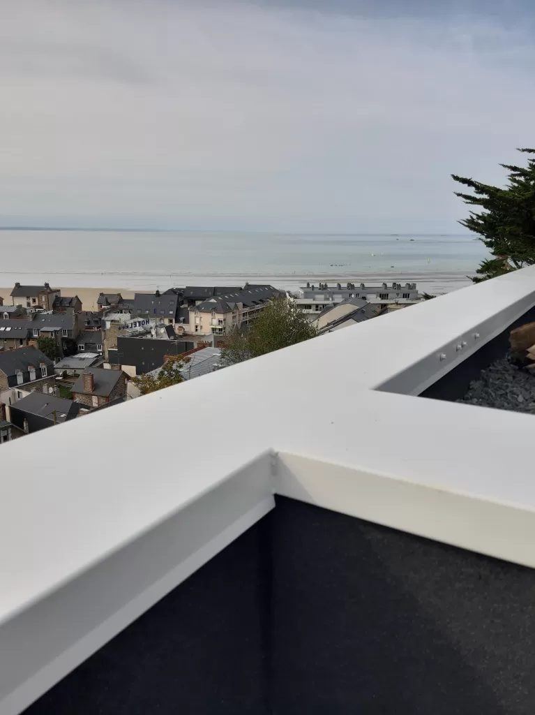 Vue d’une couvertine métallique blanche fabriquée par COMAT, installée sur un toit-terrasse avec une vue panoramique sur la mer et les toits d’une ville côtière.